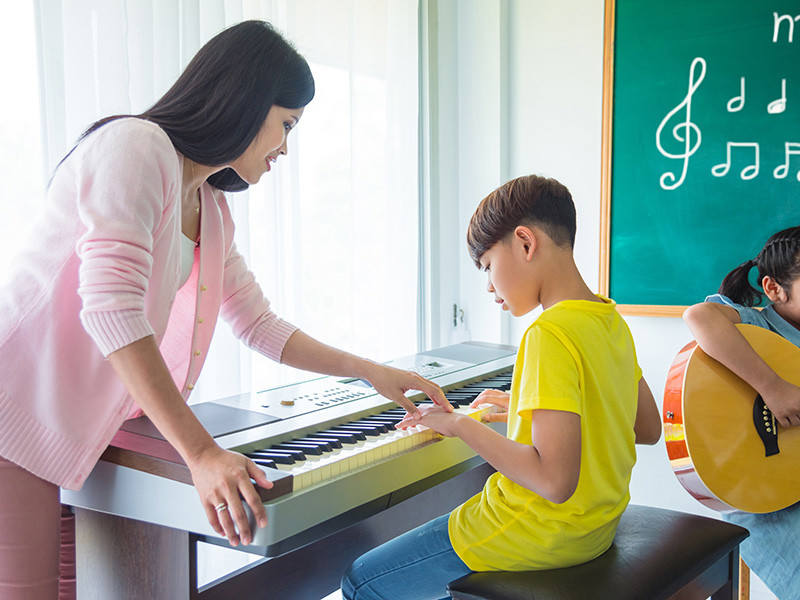 Photo of Music Therapist, Priya, who is helping a young boy on the piano